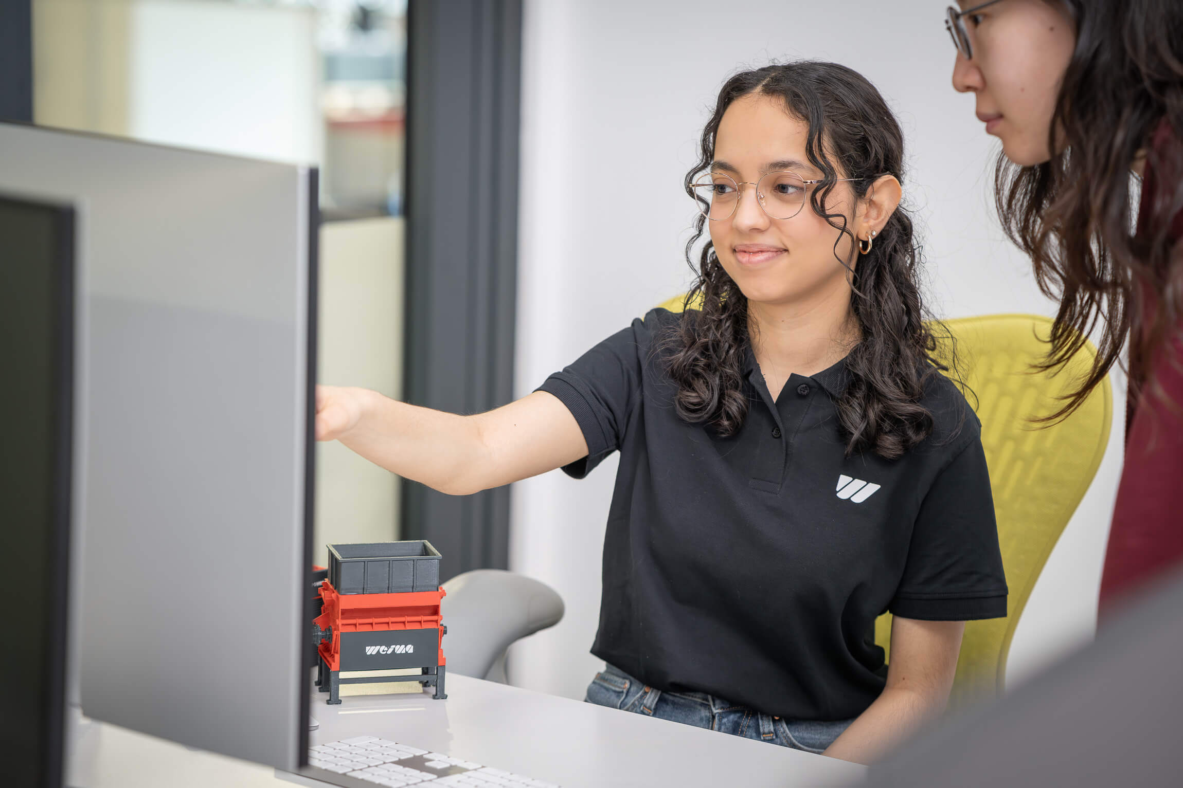 woman pointign at something in front of acomputer, 3d printed machine model next to her on a desk