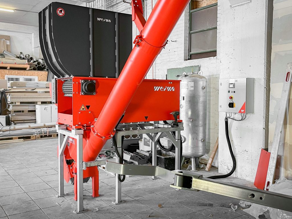 An orange shredder on a metal frame in a warehouse with a screw conveyor underneath.
