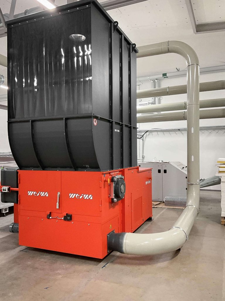 An orange shredder with a raised hopper in a warehouse with a connected extraction system.