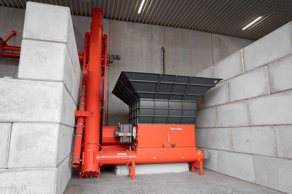An orange shredder in the middle of large concrete blocks with screw conveyor
