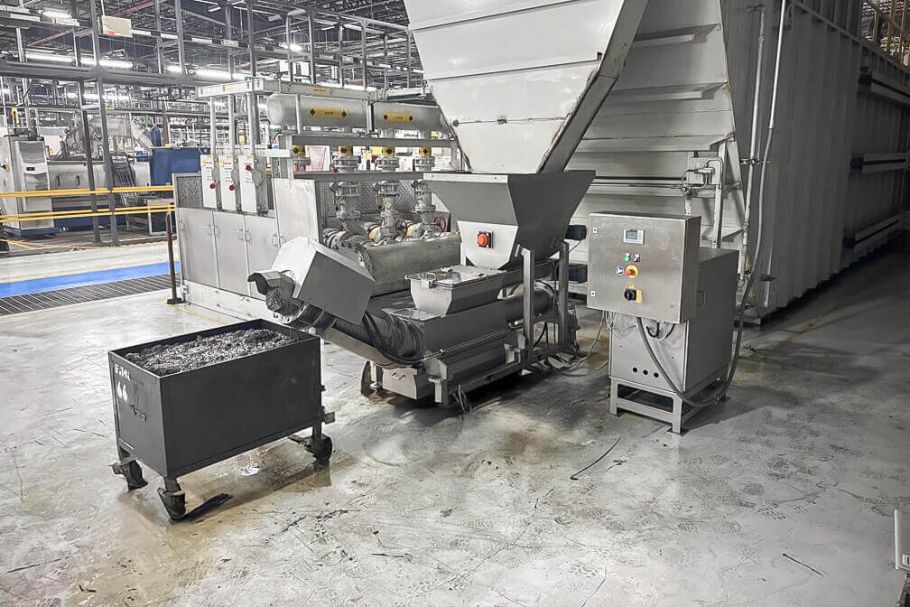 A gray drainage press in a warehouse with a full container of dry grinding sludge in front of it.