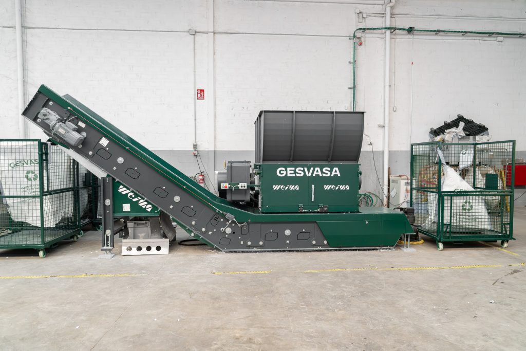 A green shredder with a conveyor belt in an industrial hall with green pallet cages