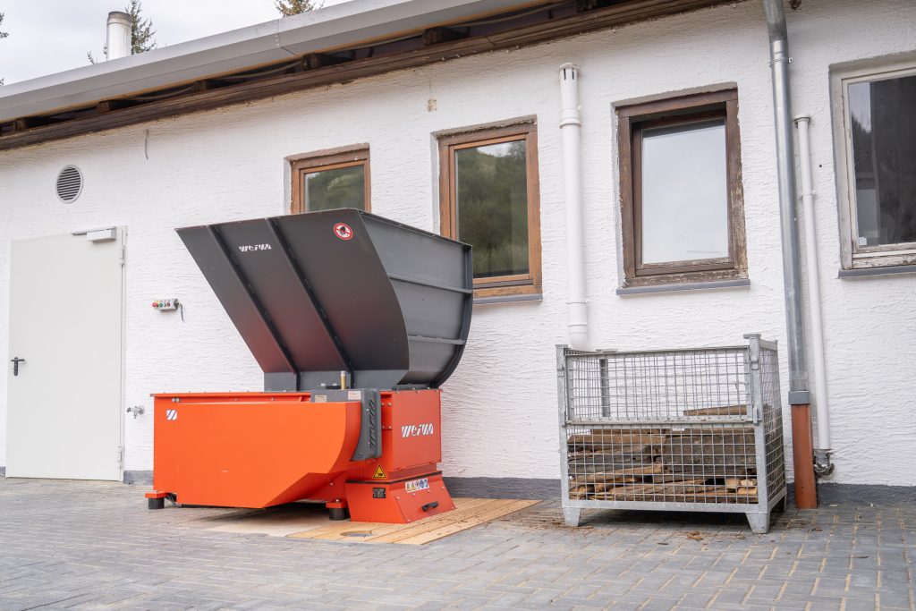 An orange shredder in front of a white building with a crate of old staves