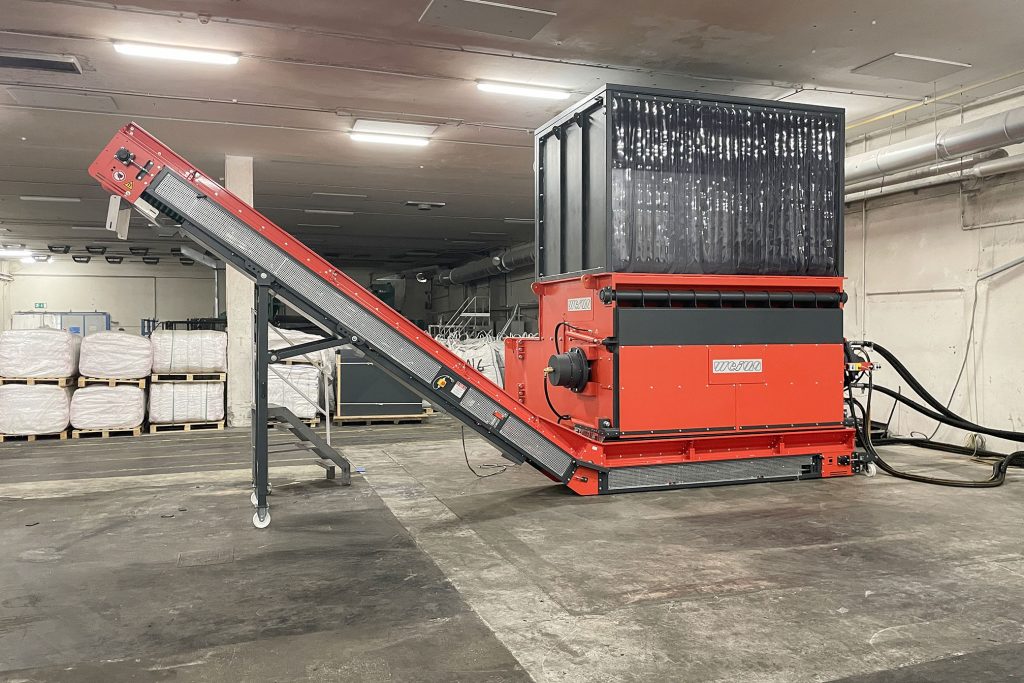An orange shredder with conveyor belt in a warehouse with material on pallets in the background