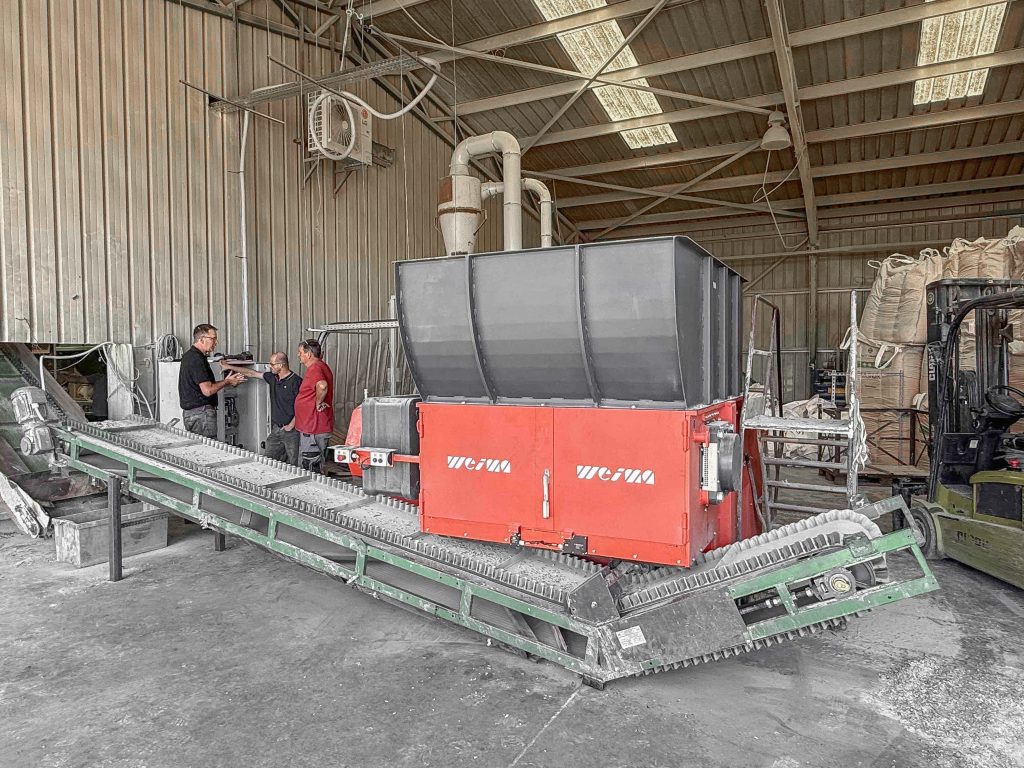 Orange shredder with a green conveyor belt in a warehouse. There are 3 men standing at the control cabinet.